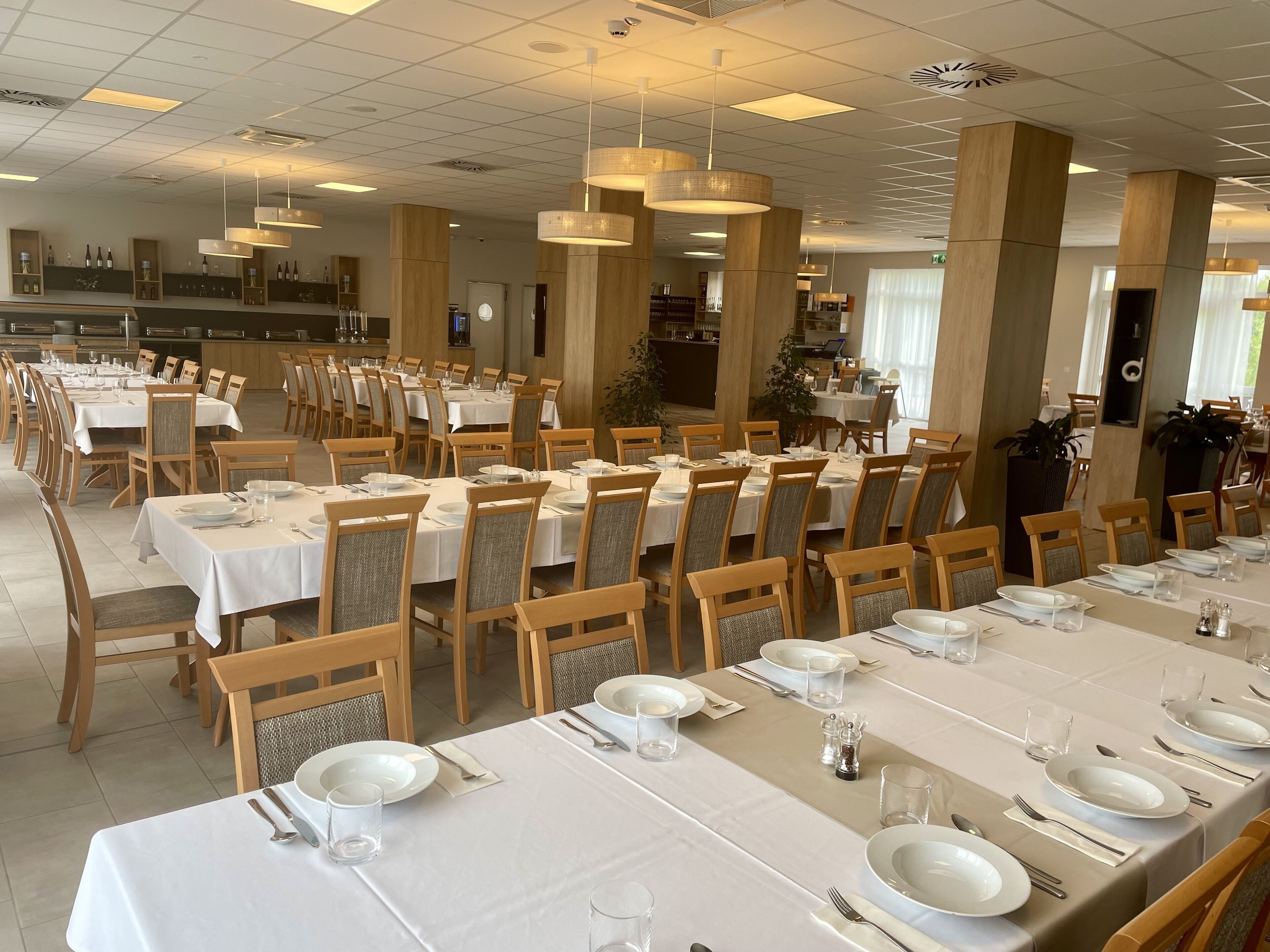 A spacious dining area with multiple tables set for service, wooden chairs, and pendant lighting.