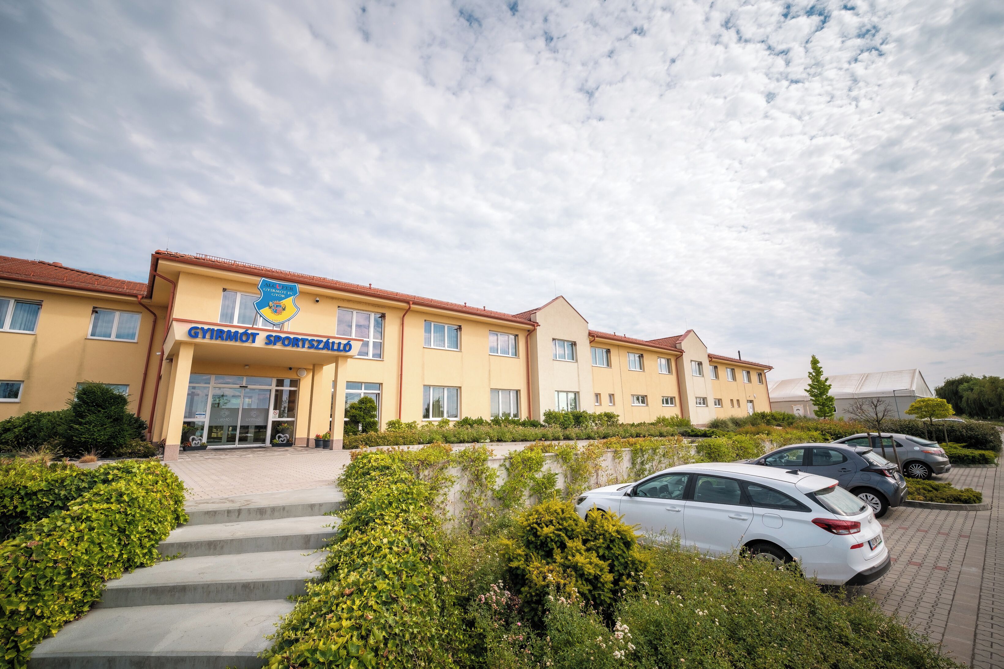 A multi-story building with a sign for 'GYIRMOT SPORTSZADE' and a parking area with cars.