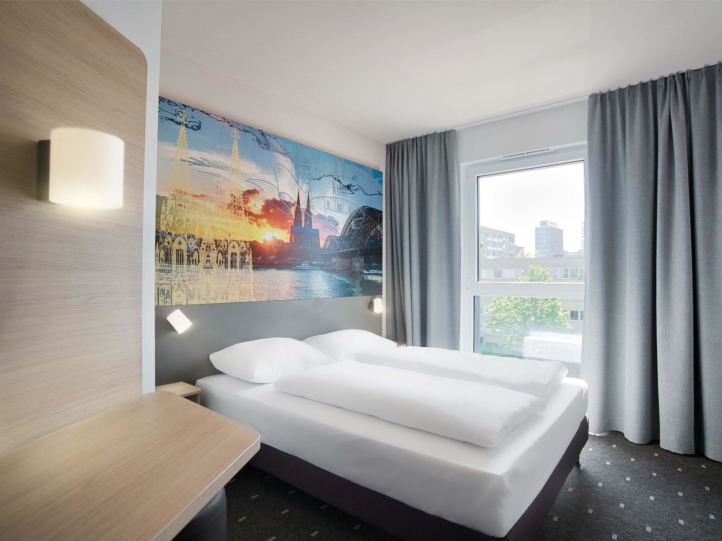 A hotel room with a bed, a painting on the wall, a window with curtains, and a view of buildings outside.