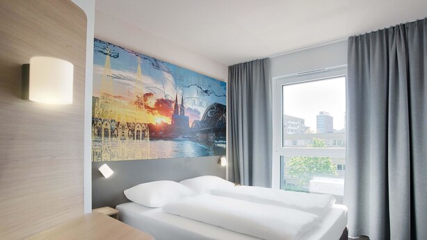 A hotel room with a bed, a painting on the wall, a window with curtains, and a view of buildings outside. - B&B Hotel Köln-City (Cologne)