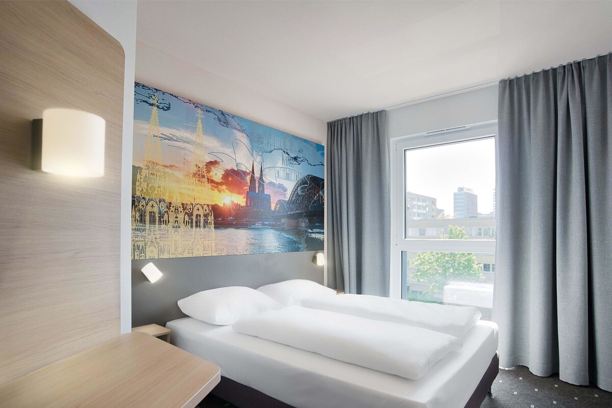 Una camera d'albergo con un letto, un quadro appeso al muro, una finestra con tende e vista sugli edifici.