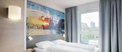 A hotel room with a bed, a painting on the wall, a window with curtains, and a view of buildings outside.