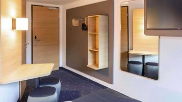 A modern hotel room with a wooden door, a small desk with a chair, a mirror, and a wall-mounted shelf.