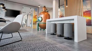 A modern interior with a large copper still, a white table with grey stools, and a flat-screen TV mounted on the wall. - B&B Hotel Köln-City (Cologne)