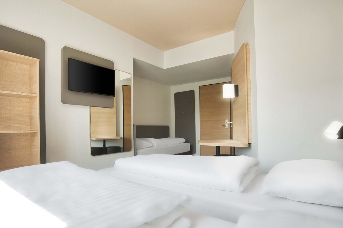 Camera d'albergo moderna con due letti, una TV a schermo piatto montata sulla parete e un comodino con lampada.