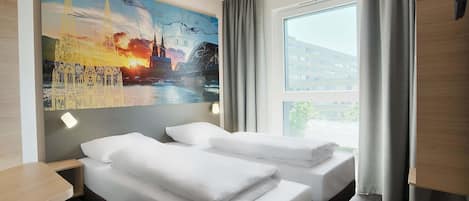 Habitación de hotel con dos camas, un ventanal con cortinas, un cuadro de un paisaje urbano y un escritorio.