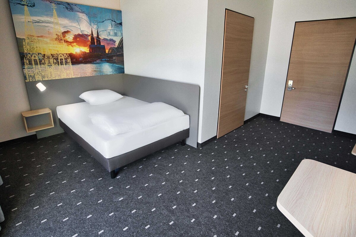 Una camera d'albergo con un letto, un comodino, una porta in legno e un quadro che raffigura un panorama urbano.