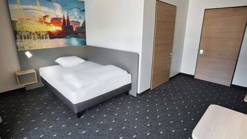 Une chambre d’hôtel avec un lit, une petite table de chevet, une porte en bois et un tableau représentant un paysage urbain accroché au mur.