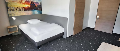 Habitación de hotel con cama, una mesita de noche pequeña, una puerta de madera y un cuadro de un paisaje urbano en la pared.