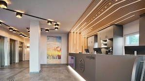 A modern reception area with a reception desk, a large poster on the wall, and a hallway with elevators. - B&B Hotel Köln-City (Cologne)