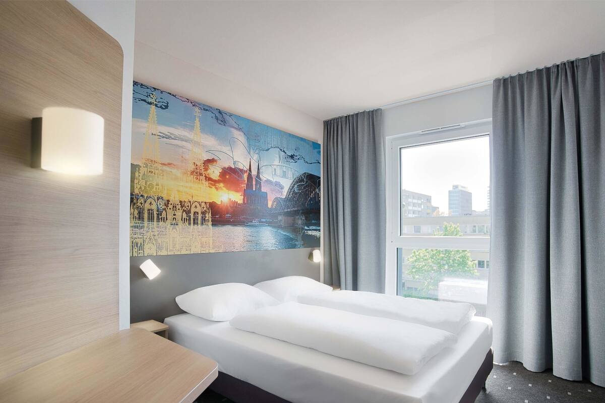 A hotel room with a bed, a painting on the wall, a window with curtains, and a view of buildings outside.