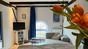 A living room with a sofa, a coffee table, a TV, and a window with blue curtains. - Landlord Doppelzimmer  22 (Kopendorf auf Fehmarn)