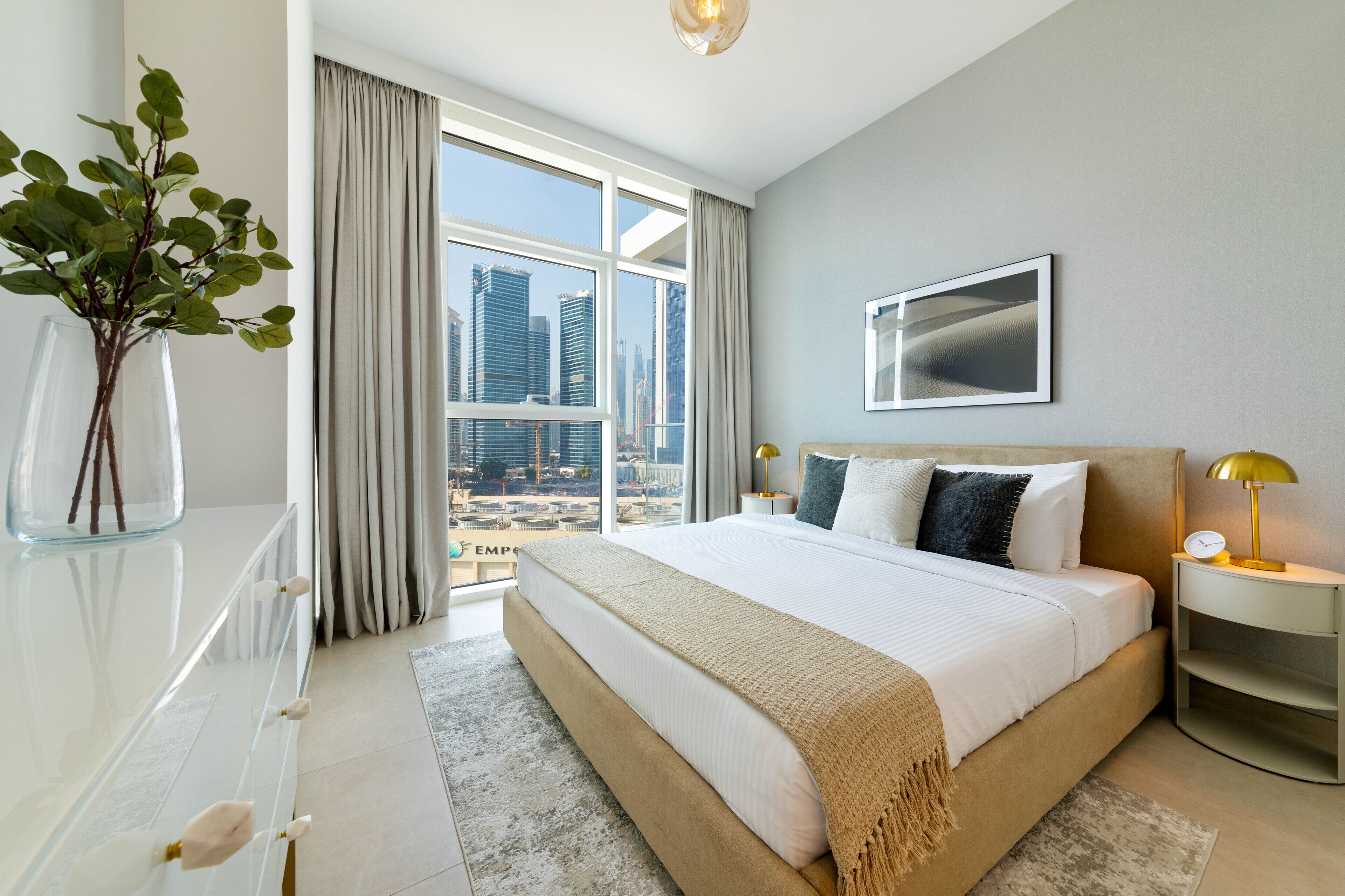 A modern hotel room with a large bed, a glass dresser, a view of the cityscape, and a cityscape painting on the wall.