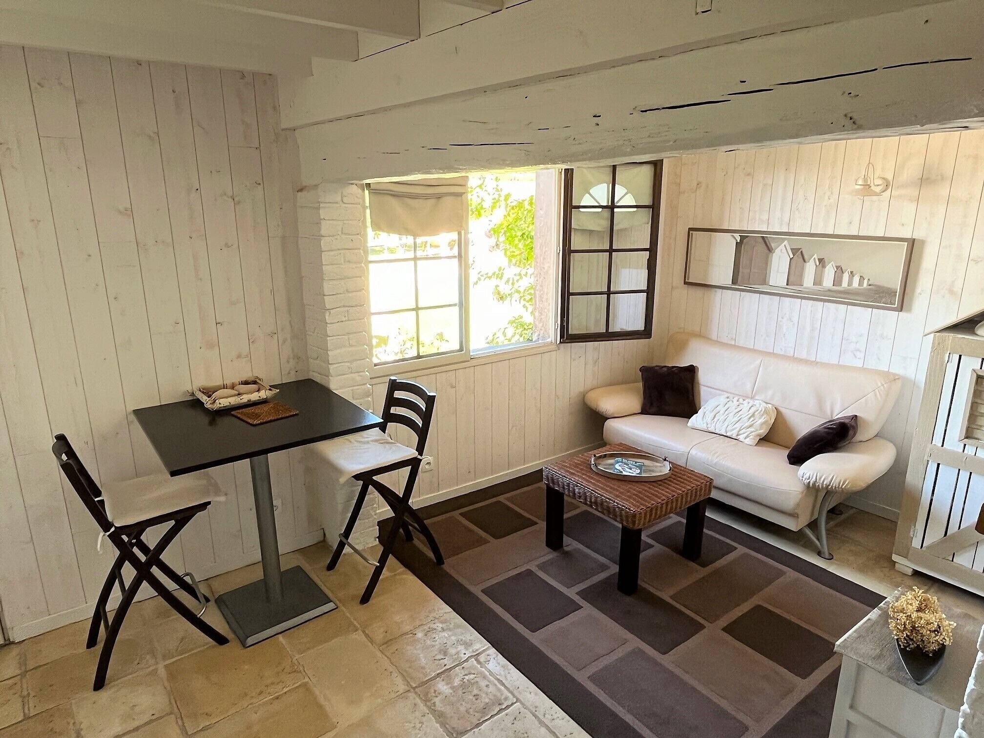 Un salon chaleureux avec un canapé blanc, une petite table et deux chaises. Une fenêtre offre une vue sur la verdure à l’extérieur.