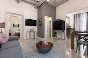 Een moderne woonkamer met een grijze bank, een salontafel, een flatscreen-tv en een eethoek met een tafel en stoelen.