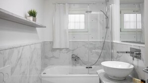 Un baño moderno con bañera, ducha con mampara de vidrio, lavamanos blanco y pared de mármol.