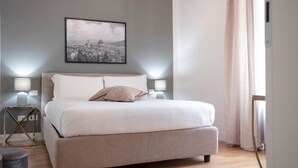 Un dormitorio con cama, lámparas de noche, un cuadro enmarco con una vista urbana y una ventana con cortinas.