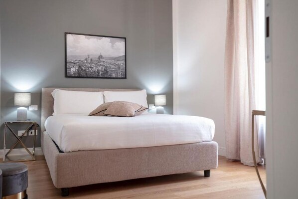 Camera da letto con un letto, lampade da comodino, una foto incorniciata di un panorama urbano e una finestra con tende.