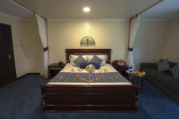 Habitación de hotel con una cama grande, una mesita de noche, un sofá y una pequeña mesa con una cesta de fruta.