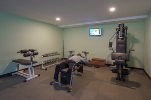 Un gimnasio con una prensa de pesas, mancuernas y un televisor en la pared.