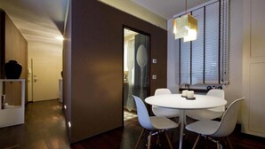 A modern dining area with a round table and white chairs, a hanging light fixture, and a window with blinds. - Luxury apartment in Ponte Vecchio (Firenze)