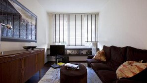 A living room with a dark brown sofa, a wooden cabinet, a television, and a large window with blinds. - Luxury apartment in Ponte Vecchio (Firenze)