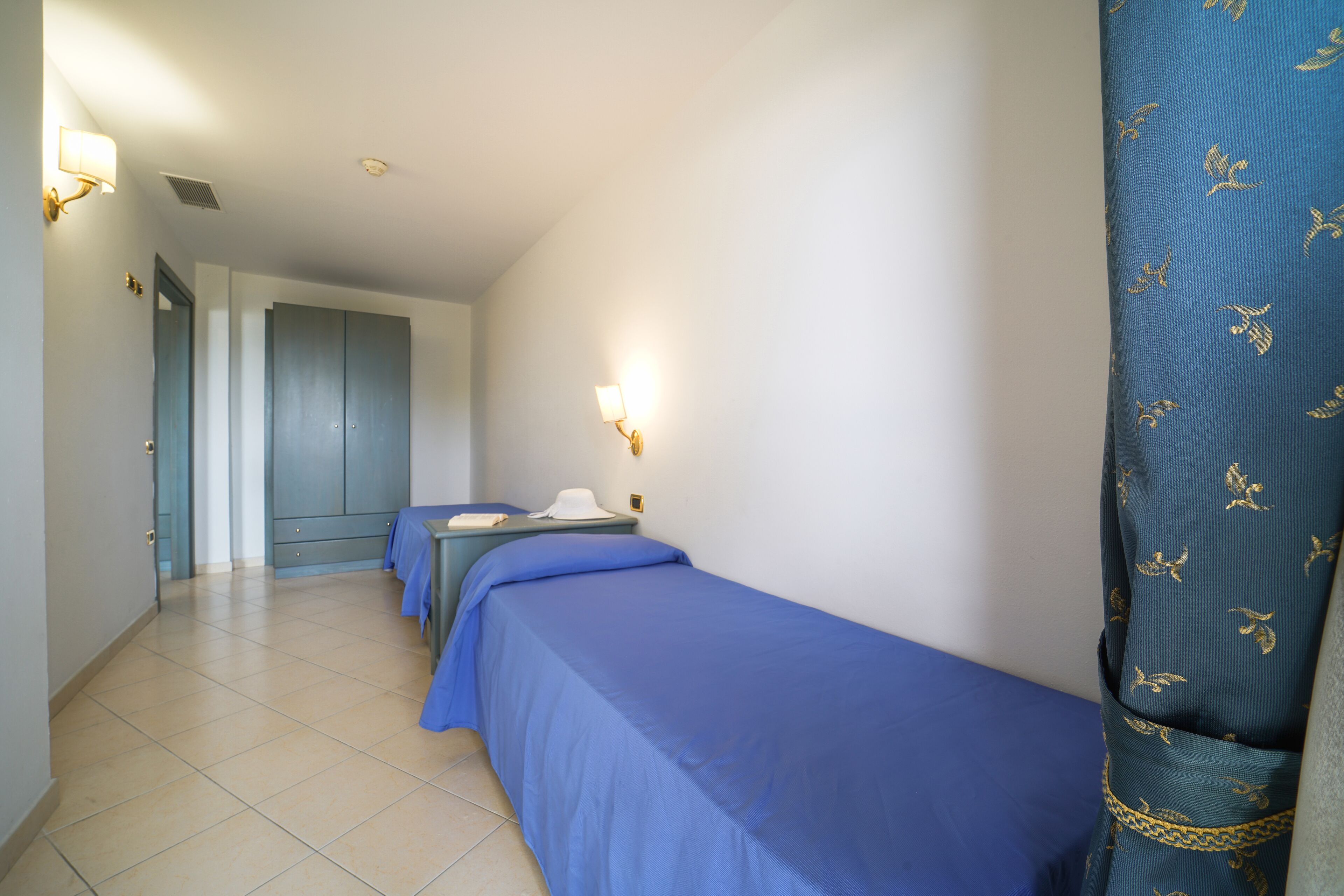Habitación de hotel con dos camas individuales, un escritorio y una silla. Se ve una puerta que da a otra habitación y una cortina con estampado de hojas.