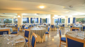 A spacious dining area with multiple round tables, blue upholstered chairs, and large windows offering views of palm trees. - Hotel Scoglio del Leone (Zambrone)