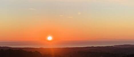 A sunset over a coastal landscape with a view of the sea and distant hills.