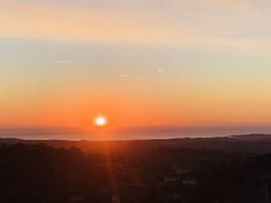 A sunset over a coastal landscape with a view of the sea and distant hills.