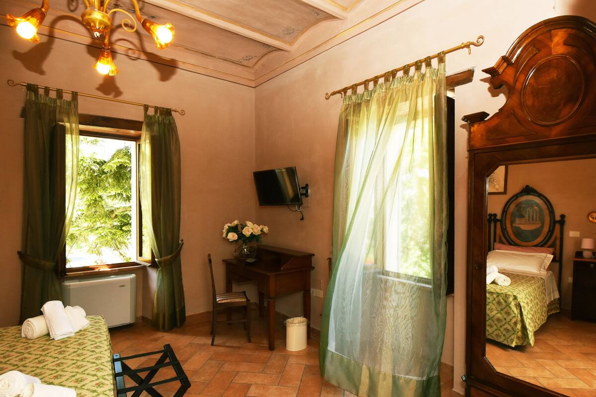 Een kamer met een bed, een bureau, een stoel, een televisie en een raam met gordijnen.