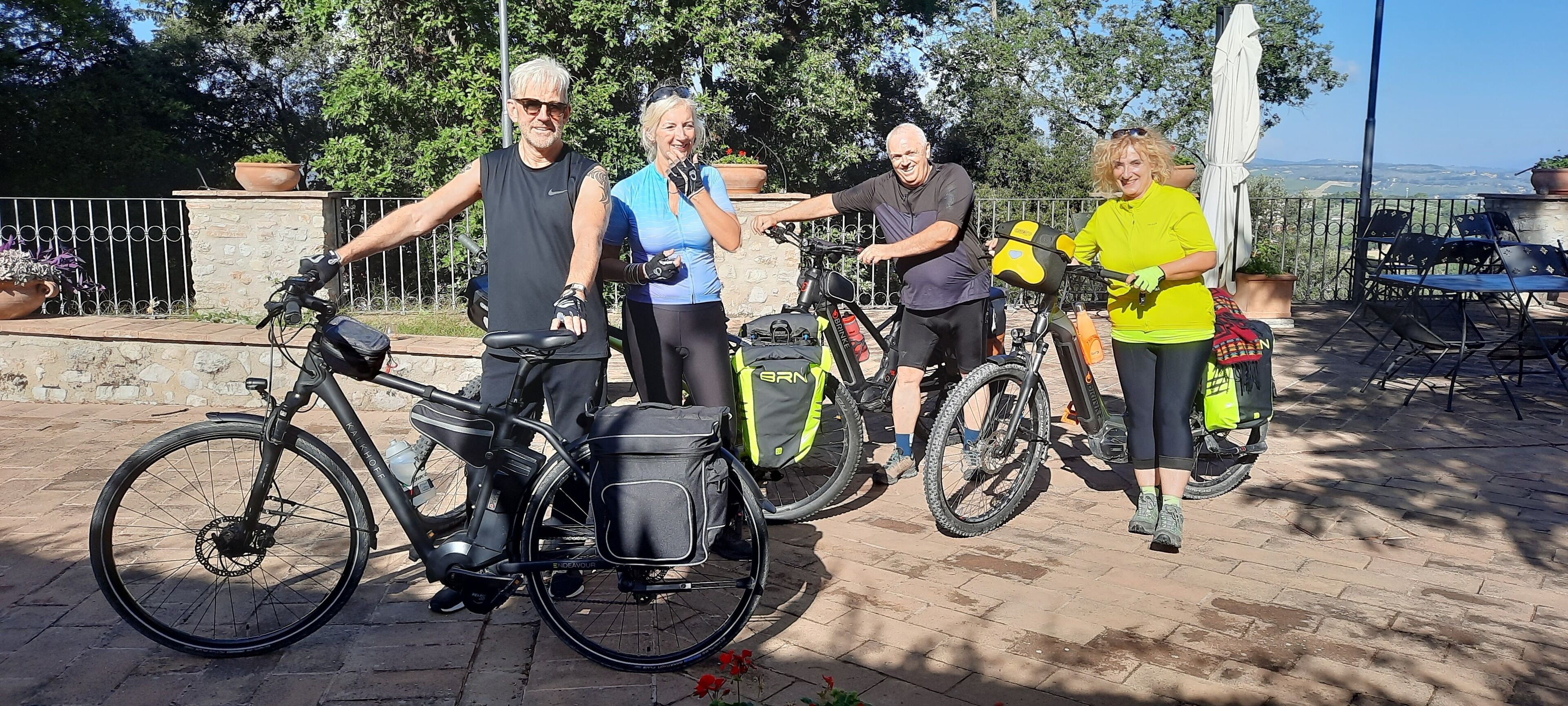 A group of people standing on a paved area with bicycles, a stone wall, and a view of the landscape in the background.