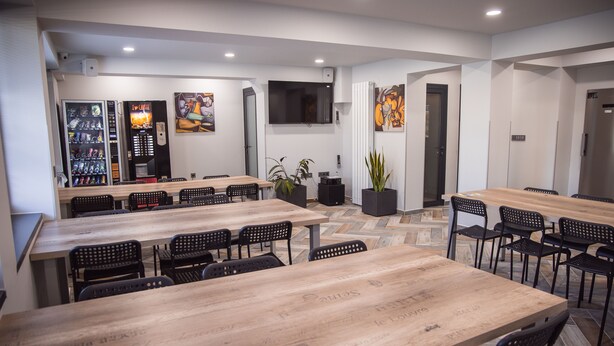 A modern dining area with long wooden tables and black chairs, a coffee machine, and a flat-screen TV mounted on the wall. - DORM BG (Stara Zagora)