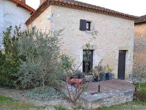 Il s’agit d’un bâtiment rustique en pierre, doté d’un toit en tuiles, d’une petite fenêtre avec des volets et d’une porte. Une zone pavée abrite des plantes en pot et une petite table.