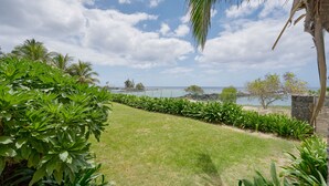 A landscaped garden with a view of the sea, featuring palm trees, a stone wall, and a grassy area. - Otera 2 Residence (Trou aux Biches)