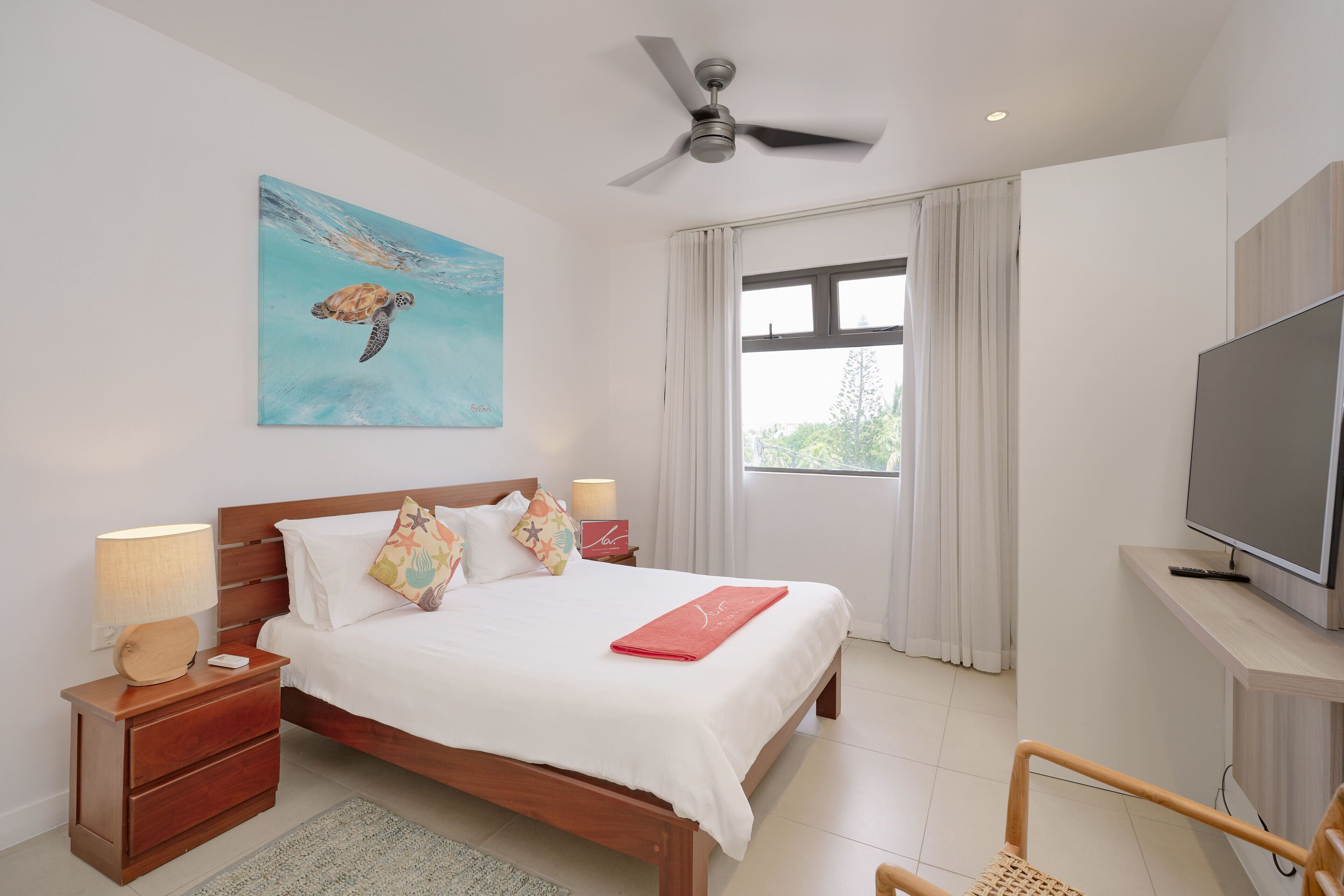 A bedroom with a bed, nightstands, a TV, a ceiling fan, and a painting of a sea turtle.