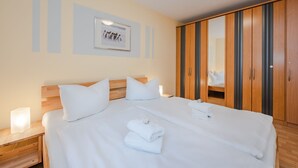 Una cama bien hecha con sábanas y almohadas blancas, una mesita de noche con lámpara y un armario de madera con espejo.
