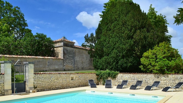 A swimming pool with lounge chairs, a stone wall, and a building with a tiled roof in the background.