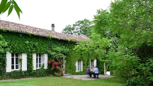 A house with ivy-covered walls and a tiled roof. There is a small garden with grass and a few flowers. Two people are sitting at a table outside.