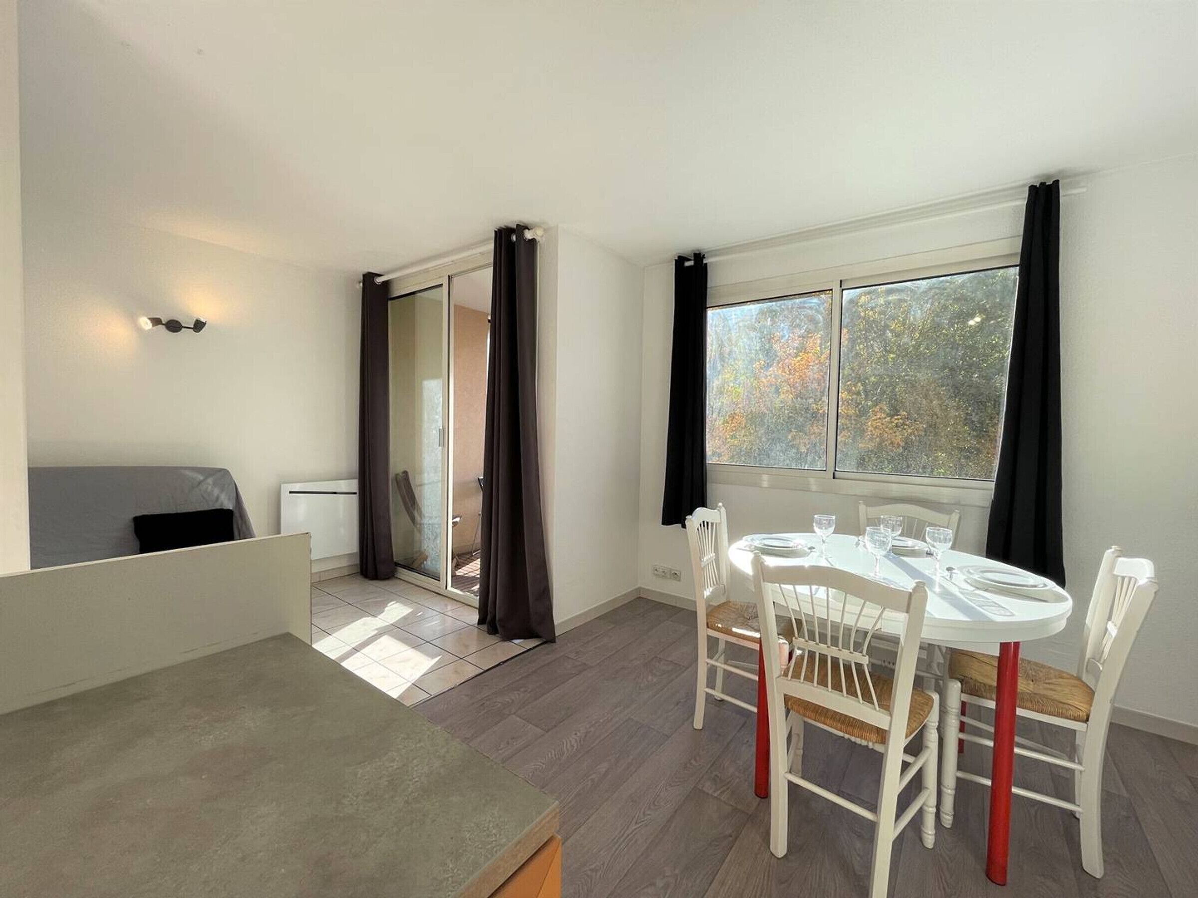 Une petite pièce lumineuse, avec une table à manger dressée pour quatre personnes, un canapé et une porte-fenêtre coulissante donnant sur un balcon.