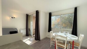 A small, bright room with a dining table set for four, a sofa, and a sliding glass door leading to a balcony.