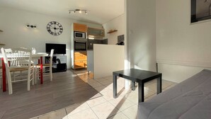 A modern living room with a grey sofa, a dining area with a white table and chairs, and a kitchen with a microwave and oven.
