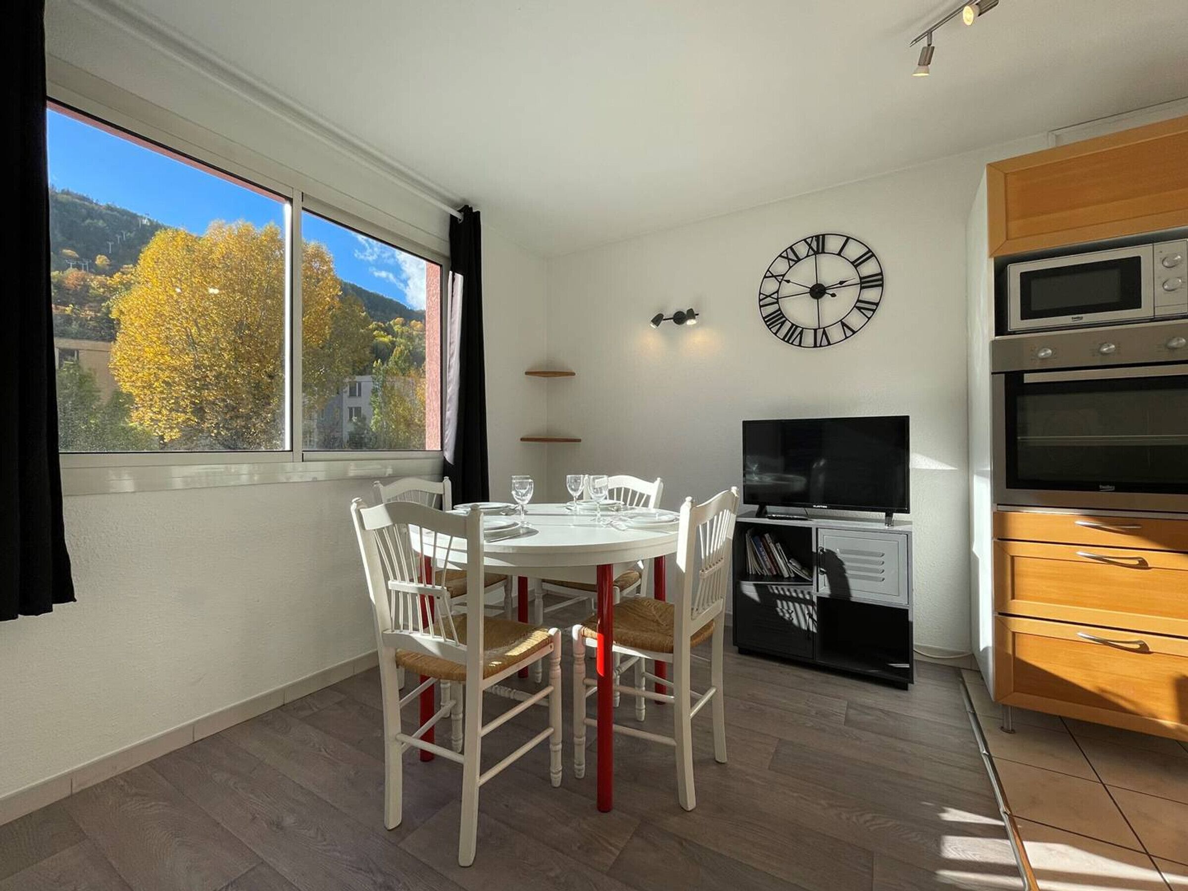 A dining area with a table and chairs, a clock on the wall, a TV, and a kitchen with appliances.