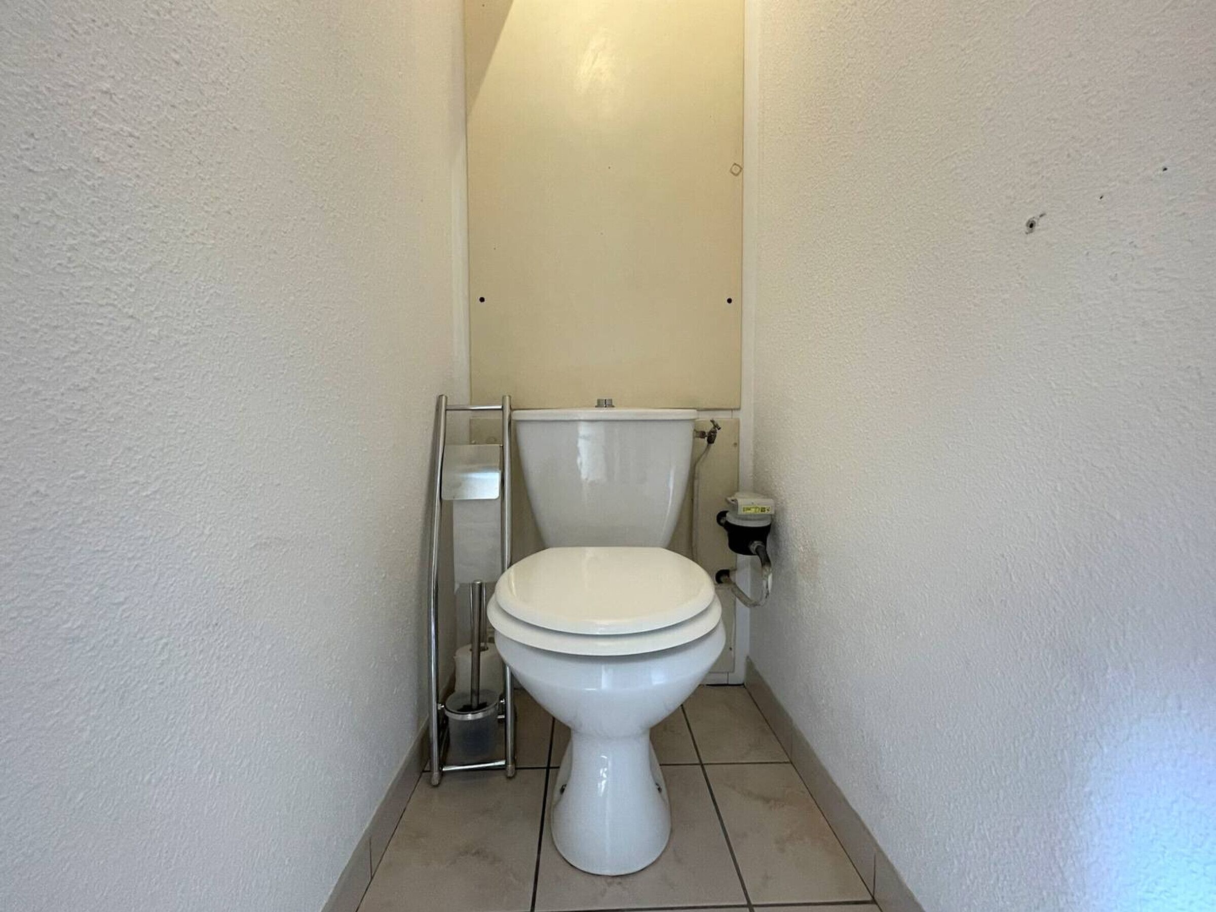 A small, narrow bathroom with a white toilet, a metal toilet paper holder, and a wall-mounted flush tank.