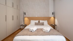 A modern hotel room with a wooden headboard, a large bed with white linens, two bedside tables, and a wall-mounted air conditioner. - Deluxe 4 Bedrooms Apartment in Athens Center (Athens)