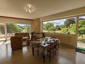 A dining area with a table set for four, surrounded by patterned sofas, large windows with a view of greenery, and a stone wall.