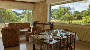 A dining area with a table set for four, surrounded by patterned sofas and chairs, with a stone wall and a large window offering a view of greenery and trees. - Comfortable house with private garden, internet, parking (Agris)
