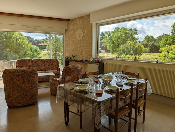 A dining area with a table set for four, surrounded by patterned sofas and chairs, with a stone wall and a large window offering a view of greenery and trees.