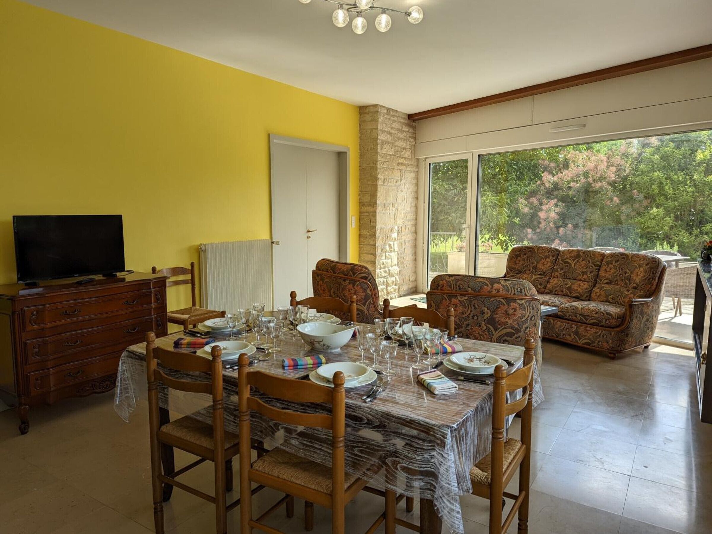 A dining area with a table set for four, surrounded by wooden chairs, a sofa, and a television on a dresser.