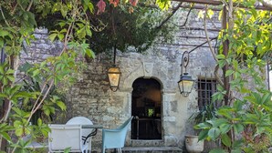 Un edificio de piedra con una entrada rústica, dos faroles colgantes y un pequeño espacio al aire libre con dos sillas.
